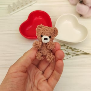 Peut inclure: Un petit ours en peluche brun avec des yeux blancs et un nez blanc est tenu dans la main d'une personne. L'ours est assis droit, les bras tendus. L'arrière-plan est une surface en bois blanc avec un plat en forme de cœur rouge et un plat en forme de cœur blanc.
