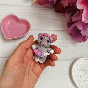 May include: A small, grey crocheted cat doll wearing a pink headband and tutu. The doll has black whiskers and white paws. A pink heart-shaped dish and pink tulips are in the background.