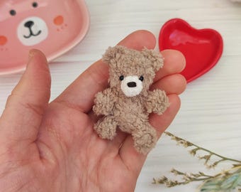 May include: A small, brown teddy bear with white embroidered eyes and nose. The bear is sitting in the palm of a hand.
