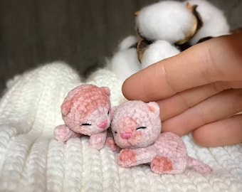 May include: Two small pink crocheted kittens lying on a white knitted blanket. The kittens have closed eyes and pink noses.