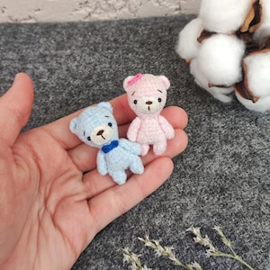 Op de afbeelding: Twee kleine gehaakte teddyberen, een blauwe en een roze, worden in een hand gehouden. De blauwe beer heeft een blauwe strik en de roze beer heeft een roze bloem op zijn hoofd.