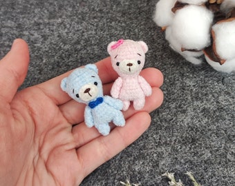 May include: Two small crocheted teddy bears, one blue and one pink, are held in a hand. The blue bear has a blue bow tie and the pink bear has a pink flower on its head.