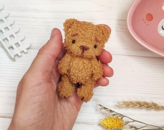 May include: A small brown teddy bear with black eyes and a brown nose is held in a hand. The bear is sitting upright with its paws together. The background is a white wooden surface with two pink heart-shaped bowls and a yellow heart-shaped bowl.