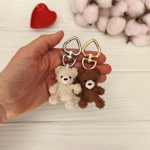 Peut inclure: Deux petits porte-clés ours en peluche, un crème et un marron, chacun attaché à un fermoir en forme de cœur. Les fermoirs sont argentés et dorés. Un petit plat rouge en forme de cœur est en arrière-plan.