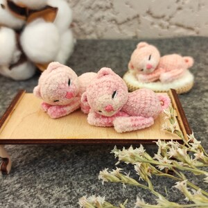 May include: Three pink crocheted cat figurines, two lying down on a wooden tray and one lying down on a white surface. The figurines have black eyes and pink noses.