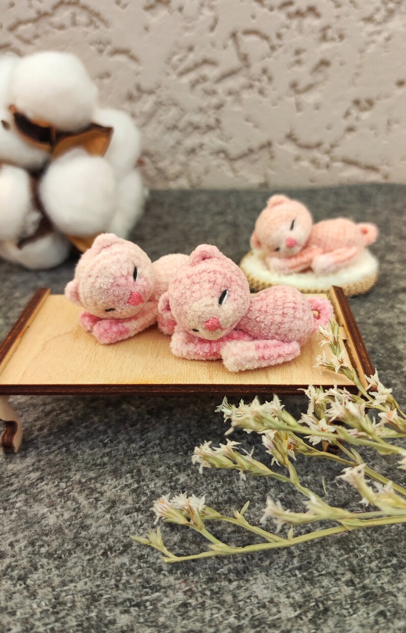 May include: Three pink crocheted cat figurines, two lying down on a wooden tray and one lying down on a white surface. The figurines have black eyes and pink noses.