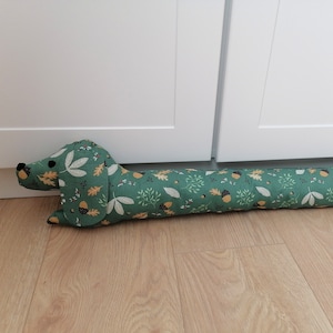 Puede incluir: Un tapón de puerta con forma de dachshund verde y marrón con un patrón de hojas y bellotas.