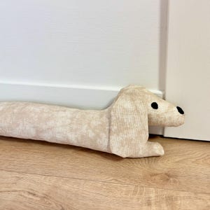 May include: A beige dachshund-shaped doorstop with black eyes. The doorstop is made of fabric and has a textured pattern.