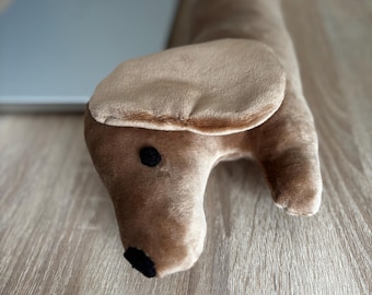 May include: A plush dachshund dog doorstop in shades of tan and brown. The dog has a long body, floppy ears, and a black nose and eyes. The doorstop is made of soft, velvety material and is designed to sit on the floor.