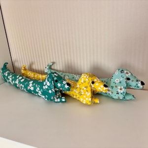 May include: Three plush dachshund dog toys in green, yellow, and light blue floral patterns. The toys are lying on a white surface.