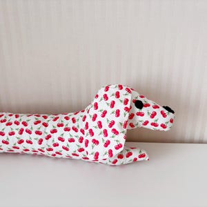 May include: A white dachshund-shaped cushion with a red cherry print. The cushion is lying on a white surface.