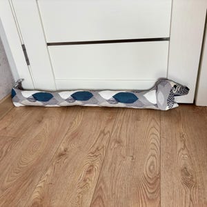 Può includere: Un fermaporta a forma di bassotto, con un motivo grigio e blu. Il fermaporta è appoggiato su un pavimento di legno.