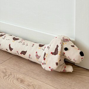 May include: A long, cylindrical fabric draft stopper shaped like a dachshund dog. The cream-colored fabric features a repeating pattern of small brown and red chickens and floral sprigs. It has two black fabric circles for eyes and a black nose.