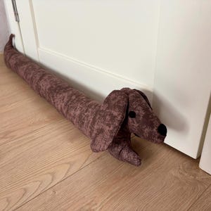 May include: A brown dachshund-shaped doorstop with a textured fabric. The doorstop is lying on a wooden floor.