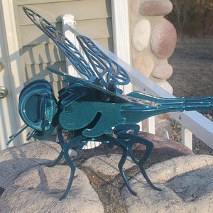 May include: Teal blue metal dragonfly sculpture with intricate details. The dragonfly is perched on a rock with its wings spread out.