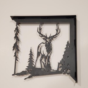 May include: Black metal wall decor featuring a silhouette of an elk standing in a forest with pine trees. The elk is facing forward with its antlers raised.