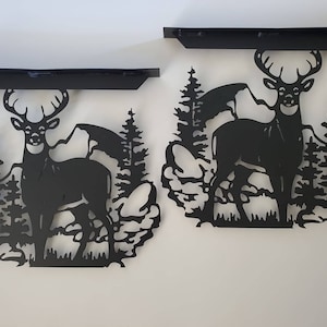 May include: Two black metal shelf brackets with a deer silhouette design. The deer is standing in a forest setting with trees and mountains in the background.