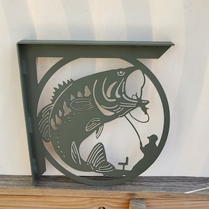 May include: A green metal shelf bracket with a cutout design of a bass fish jumping out of the water. The fish is detailed with scales and fins. A silhouette of a person fishing is visible in the background.