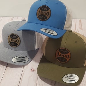 May include: Three trucker hats with different colored brims and mesh back panels. Each hat features a brown leather patch with the text "YOUR LOGO HERE" in black.