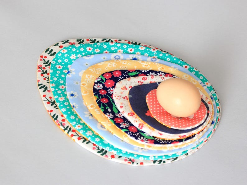 Puede incluir: Un conjunto de siete c&iacute;rculos de tela, cada uno con un patr&oacute;n floral diferente, dispuestos en un patr&oacute;n de c&iacute;rculo conc&eacute;ntrico. Los c&iacute;rculos son en tonos de azul, amarillo, rojo y blanco. Un huevo beige est&aacute; en el centro de los c&iacute;rculos.