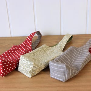 May include: Three fabric wine bottle bags. One is red with white polka dots, one is beige with a brown and white pattern, and one is gray with white stripes.
