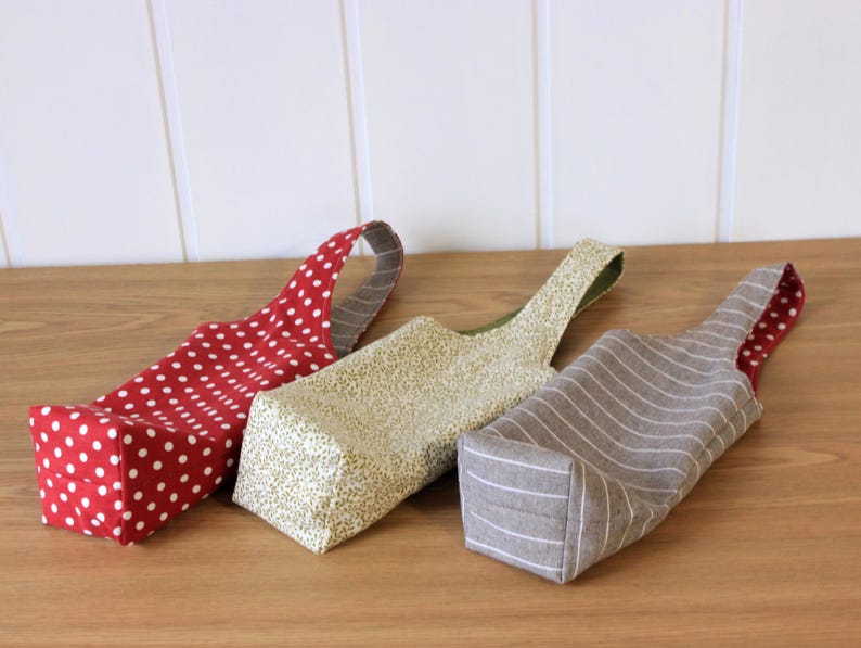 May include: Three fabric wine bottle bags. One is red with white polka dots, one is beige with a brown and white pattern, and one is gray with white stripes.