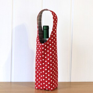 May include: Red wine bottle carrier with white polka dots. The carrier has a handle and is made of fabric.