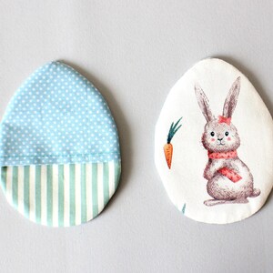 Puede incluir: Dos adornos de Pascua en forma de huevo de tela. Uno es azul con lunares blancos y un fondo a rayas verde y blanco. El otro es blanco con un conejito marr&oacute;n que lleva una bufanda roja y una zanahoria.