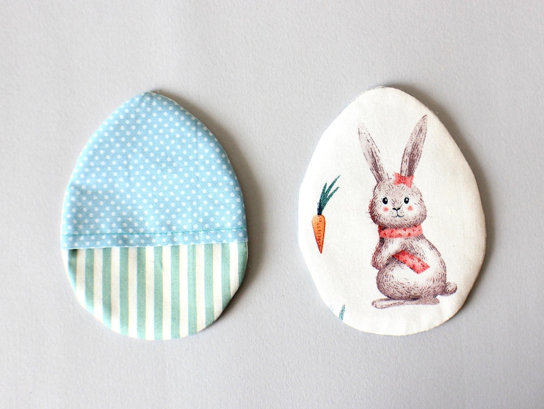 Puede incluir: Dos adornos de Pascua en forma de huevo de tela. Uno es azul con lunares blancos y un fondo a rayas verde y blanco. El otro es blanco con un conejito marr&oacute;n que lleva una bufanda roja y una zanahoria.