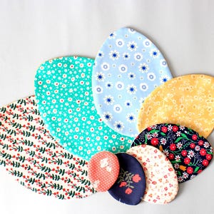 Puede incluir: Un conjunto de siete posavasos de tela con forma de huevo en varios patrones florales, incluyendo azul, amarillo, verde, rojo y blanco.
