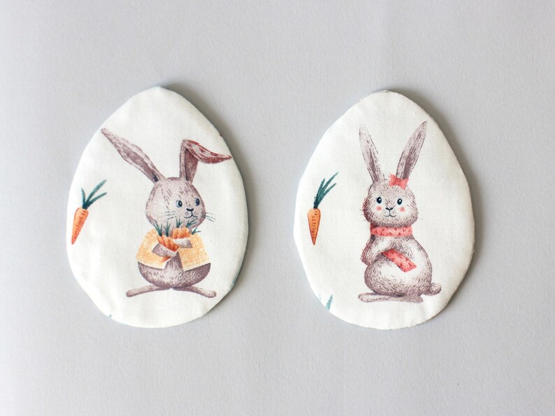 Puede incluir: Dos huevos de Pascua de tela de forma ovalada blanca con ilustraciones de conejos. Un conejo lleva una chaqueta amarilla y sostiene zanahorias. El otro conejo lleva una bufanda rosa y sostiene una zanahoria.