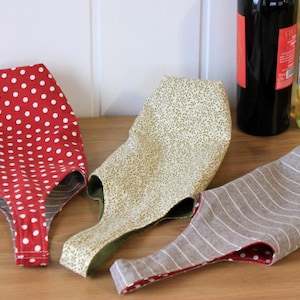 May include: Three fabric wine bottle carriers. One is red with white polka dots, one is beige with a floral pattern, and one is gray with white stripes.