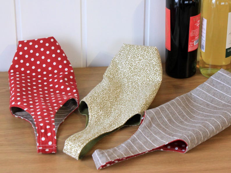 May include: Three fabric wine bottle carriers. One is red with white polka dots, one is beige with a floral pattern, and one is gray with white stripes.