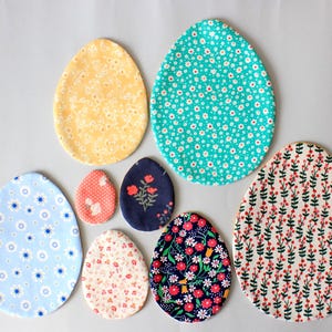 Puede incluir: Siete huevos de Pascua de tela en varios patrones y colores. Los huevos est&aacute;n hechos de tela y tienen una variedad de dise&ntilde;os florales y geom&eacute;tricos. Los huevos est&aacute;n dispuestos en un semic&iacute;rculo sobre un fondo blanco.