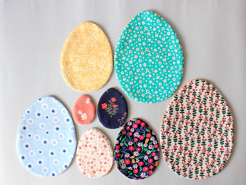 Puede incluir: Siete huevos de Pascua de tela en varios patrones y colores. Los huevos est&aacute;n hechos de tela y tienen una variedad de dise&ntilde;os florales y geom&eacute;tricos. Los huevos est&aacute;n dispuestos en un semic&iacute;rculo sobre un fondo blanco.