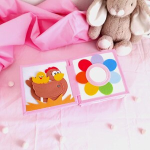 May include: A soft, open book with felt illustrations of a hen and chick, and a colorful flower. The book is pink and white, and is on a pink surface. A stuffed bunny is in the background.