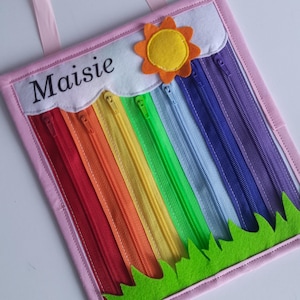 May include: A pink felt board with a rainbow of zips, a yellow sun with orange rays, and green grass. The board is personalised with the name "Maisie".