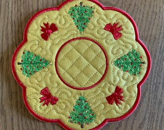Op de afbeelding: Een gele gestikte onderzetter met een rode rand en vijf geborduurde kerstboompjes. De boompjes zijn groen met rode strikken en glitteraccenten.