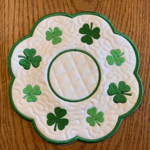 Puede incluir: Un posavasos acolchado blanco con un borde bordado verde y diseños de tréboles. El posavasos tiene un centro redondo para una taza o taza.
