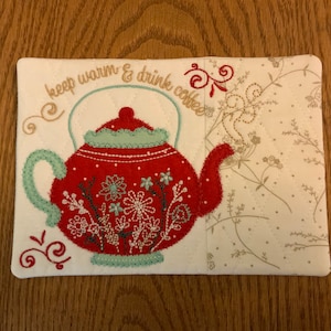 May include: A white fabric tea cozy with embroidered red teapot, green leaves, and the text "keep warm & drink coffee".