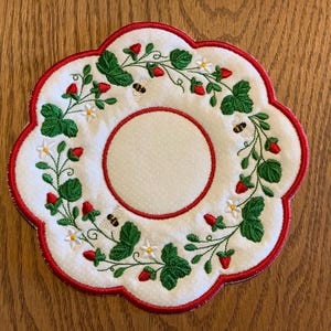 May include: A white circular fabric coaster with a scalloped edge trimmed in red thread. The design features an embroidered wreath of green strawberry leaves, small red strawberries, white flowers, and three small bees circling the center.