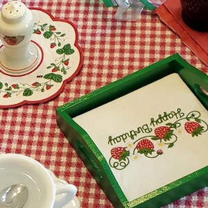 May include: A square green wooden tray holding a white napkin printed with red strawberries and the text Happy Birthday in green script. Nearby is a white ceramic salt shaker on a matching scalloped plate with strawberry motifs and a white cup.