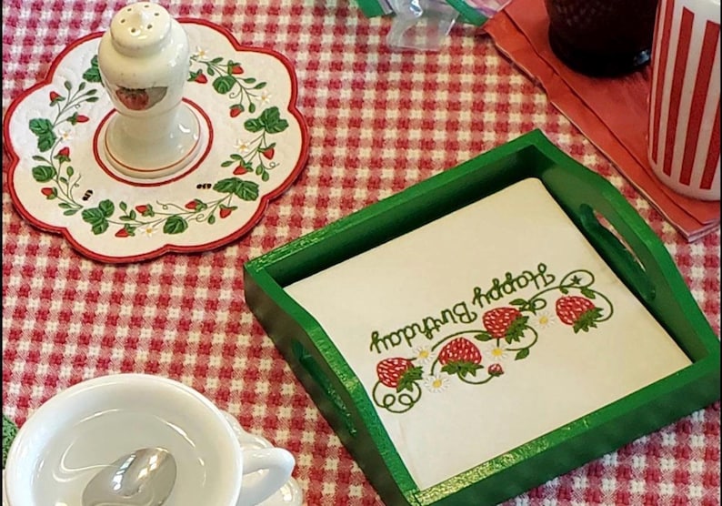 May include: A square green wooden tray holding a white napkin printed with red strawberries and the text Happy Birthday in green script. Nearby is a white ceramic salt shaker on a matching scalloped plate with strawberry motifs and a white cup.