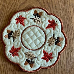 May include: A round, quilted coaster with a brown scalloped edge. The coaster is decorated with embroidered autumn leaves in shades of red, orange, and brown, and acorns in brown. The center of the coaster is a circle of quilted fabric.