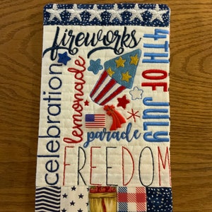 May include: A rectangular, quilted fabric item with patriotic embroidery. The design features the words "Fireworks," "Celebration," "Lemonade," "Freedom," and "4th of July." It includes images of a firework, stars, and the American flag. The edges are trimmed with a blue and white star pattern.
