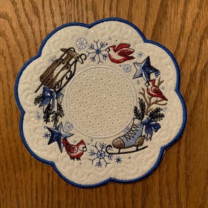May include: White round coaster with a blue scalloped edge. The coaster features an embroidered design of a winter scene with red cardinals, a blue ice skate, a star, and a sled.