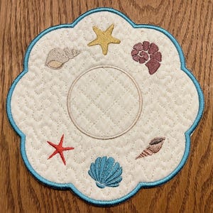 May include: A white quilted coaster with a blue scalloped edge features embroidered seashells, starfish, and a circle in the center.