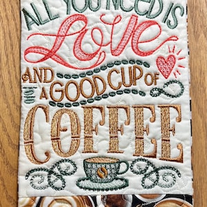 May include: Embroidered coffee-themed wall hanging with the text "ALL YOU NEED IS LOVE AND A GOOD CUP OF COFFEE." Features a coffee cup design and a brown and white patterned border. The background is white.