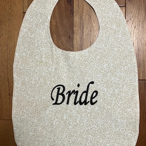Puede incluir: Un babero color crema con un patr&oacute;n de remolinos repetidos. La palabra "Bride" est&aacute; bordada en escritura negra en la parte delantera. El babero tiene una forma curva con una abertura para el cuello y un cierre de velcro.