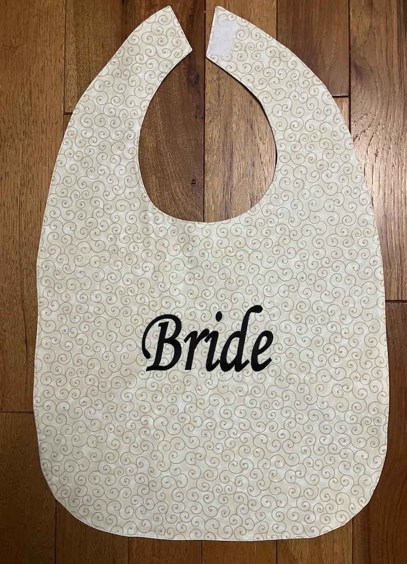 Puede incluir: Un babero color crema con un patr&oacute;n de remolinos repetidos. La palabra "Bride" est&aacute; bordada en escritura negra en la parte delantera. El babero tiene una forma curva con una abertura para el cuello y un cierre de velcro.